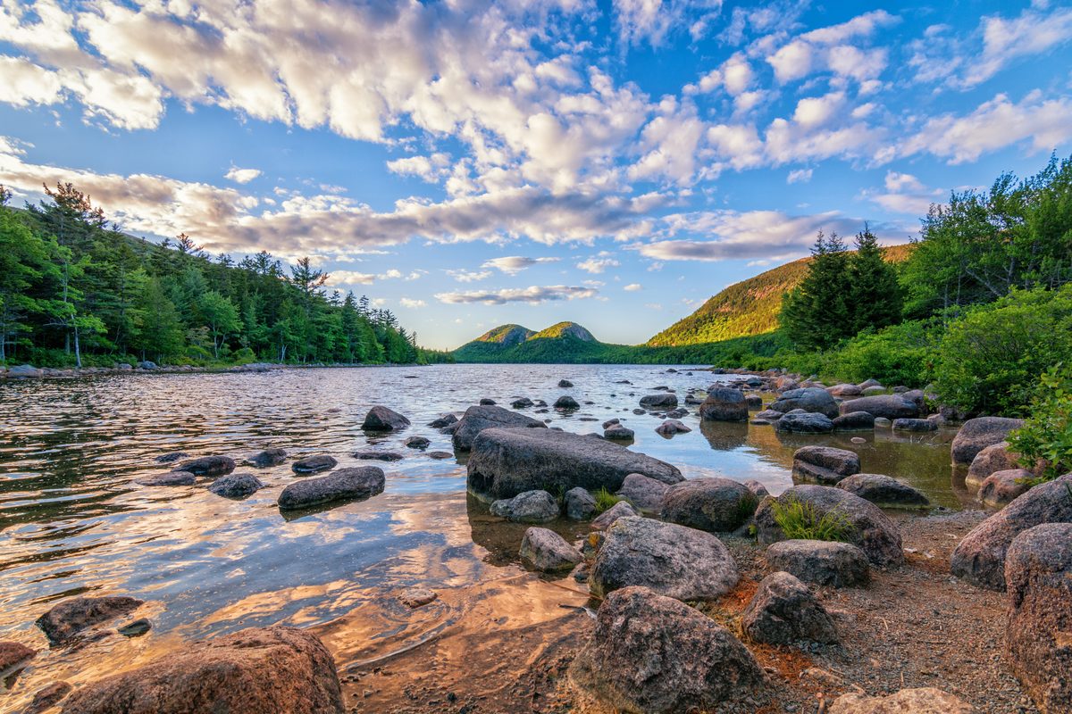 Jordan Pond in Acadia National Park, Maine, featuring smooth glacial boulders along the shoreline with the iconic Bubbles mountains rising in the background, bathed in warm golden-hour light against a dramatic blue sky filled with altocumulus clouds.