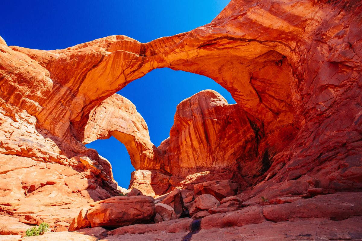 Double Arch at Arches National Park in Utah, two natural sandstone arches framing a vivid cobalt blue sky, photographed from a dramatic low-angle perspective that emphasizes the towering scale and warm orange-red hues of the Entrada sandstone.