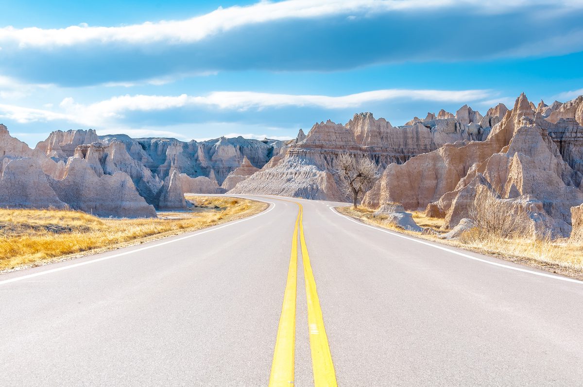 A two-lane highway with double yellow center lines winds through the dramatic eroded buttes and spires of Badlands National Park, South Dakota, under a partly cloudy blue sky. The scene has an open, inviting mood with warm golden grasses flanking…