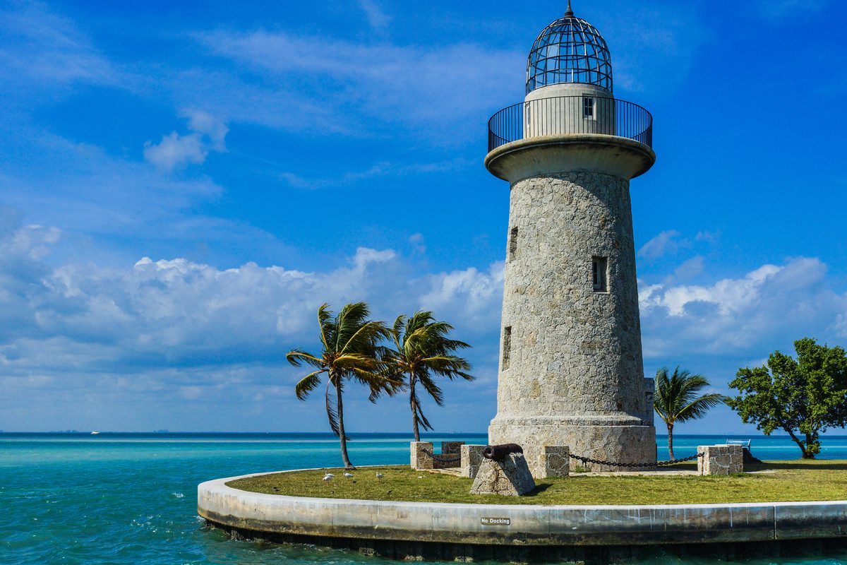 The Boca Chita Key lighthouse in Biscayne National Park stands on a small coral island surrounded by turquoise waters, with palm trees swaying in the breeze under a vibrant blue sky with scattered clouds. The historic stone tower is bathed in bright…