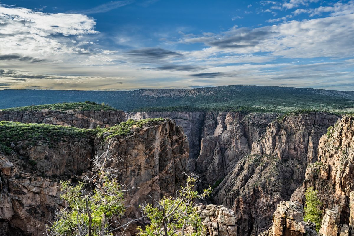 A dramatic aerial view of the Black Canyon of the Gunnison, featuring sheer dark metamorphic rock walls plunging into a deep narrow gorge, under a partially cloudy blue sky with warm afternoon light illuminating the canyon rim.
