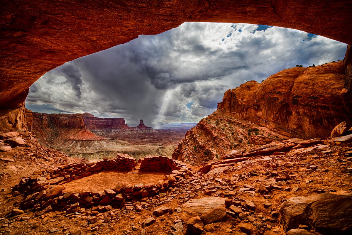 A dramatic view from inside a natural sandstone alcove at False Kiva in Canyonlands National Park, revealing ancient stone ruins in the foreground and sweeping canyon mesas under a stormy, cloud-laden sky. The warm red-orange tones of the rock…