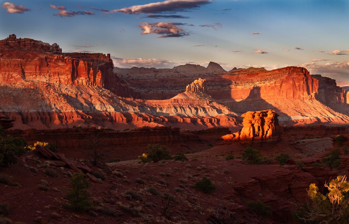A sweeping panorama of red sandstone mesas and layered canyon walls bathed in warm golden-hour light, likely Capitol Reef National Park in Utah, with dramatic shadows and glowing orange cliffs set against a blue sky with scattered clouds.