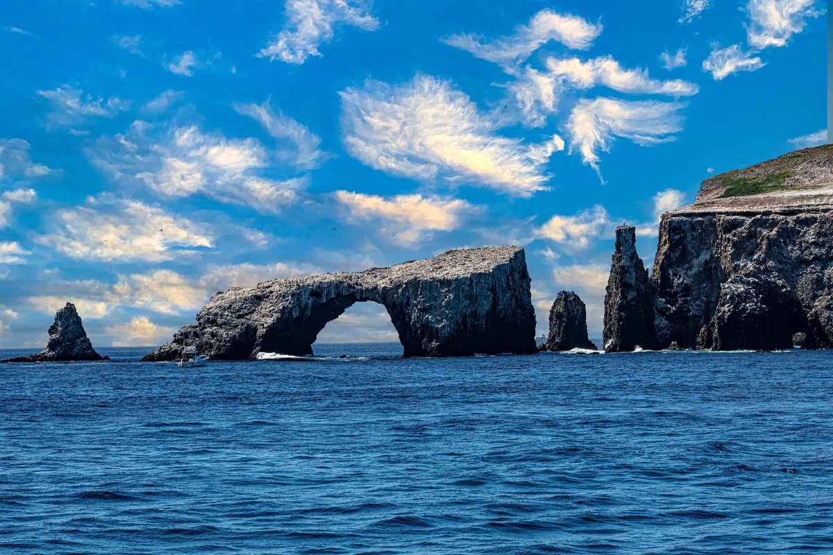 Anacapa Island's iconic Arch Rock in Channel Islands National Park, California — a weathered volcanic sea-arch rises from a deep blue Pacific alongside a slender rock spire, with sandstone island bluffs flanking the right and high cirrus clouds streaking a vivid summer sky.