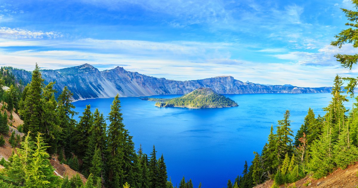 A sweeping panoramic view of Crater Lake National Park in Oregon, featuring the iconic deep cobalt-blue caldera lake with Wizard Island rising from its center, framed by evergreen conifers and dramatic volcanic crater walls under a bright blue…