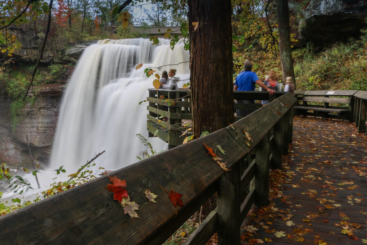 A powerful waterfall cascades over layered rock ledges into a misty gorge, viewed from a wooden observation deck surrounded by autumn foliage in warm oranges, reds, and greens. The long-exposure silky water contrasts beautifully with the rustic…