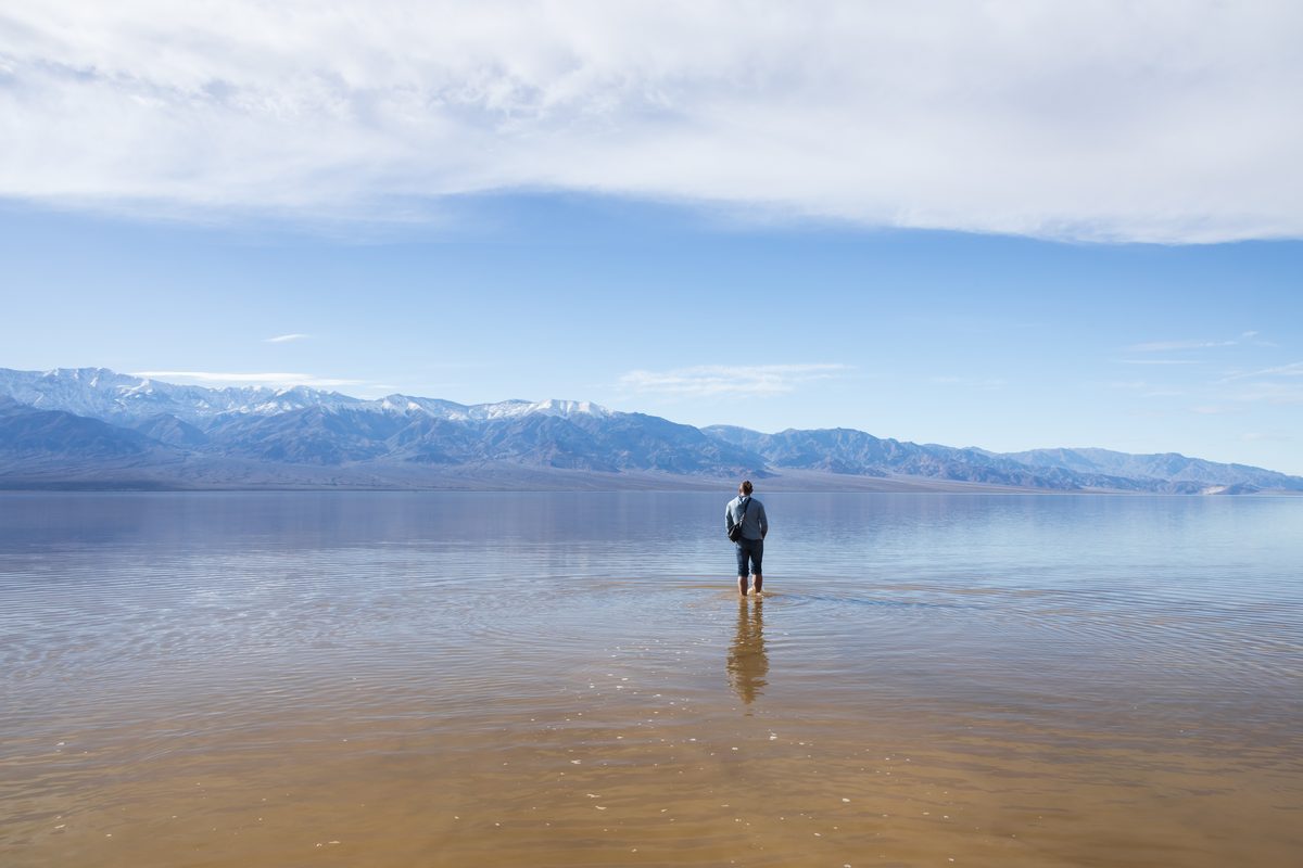 A lone figure wades through the shallow, mirror-like waters of a flooded desert basin — likely Badwater Basin in Death Valley National Park — with snow-dusted mountain ranges stretching across the horizon under a expansive blue sky with wispy…