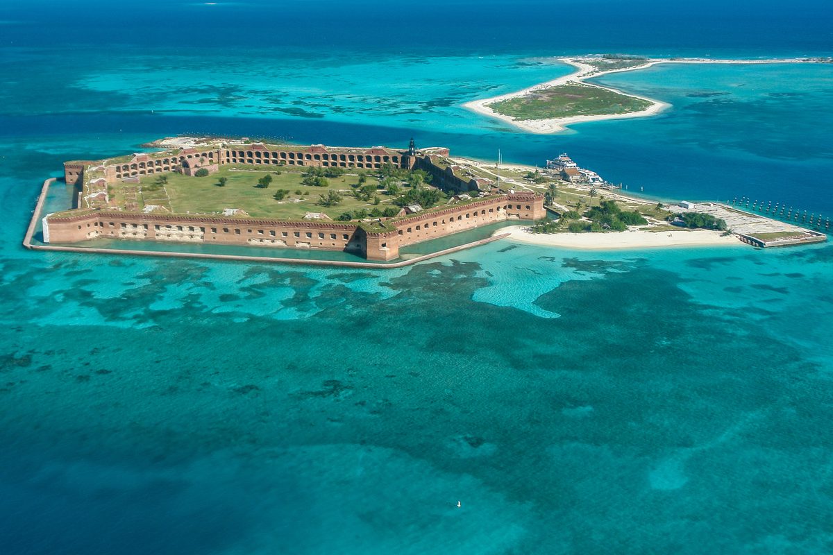 An aerial view of Fort Jefferson on Garden Key in Dry Tortugas National Park, Florida, surrounded by vivid turquoise and teal shallow waters with visible coral reefs. The massive hexagonal 19th-century brick fort sits on a small island under bright…