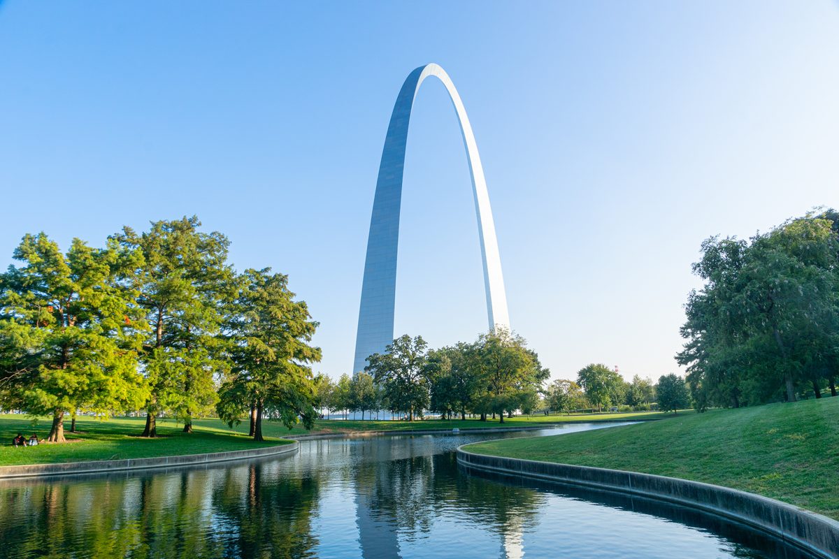 The iconic Gateway Arch in St. Louis rises dramatically against a clear blue sky, reflected in a calm reflecting pool surrounded by lush green parkland. Warm morning light illuminates the stainless steel structure and verdant trees, creating a…