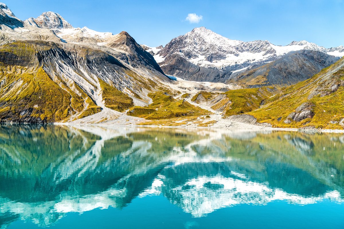 A stunning alpine lake with vivid turquoise water perfectly mirrors snow-capped mountain peaks and glaciers in calm, glassy conditions. Bright midday light illuminates golden-green tundra slopes, rocky moraines, and dramatic glaciated summits under…