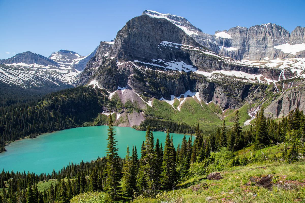 A stunning alpine lake with vivid turquoise glacial water nestled among dense evergreen forests and dramatic snow-dusted rocky peaks, photographed under bright midday sunlight with crisp blue skies conveying a sense of grandeur and wilderness.