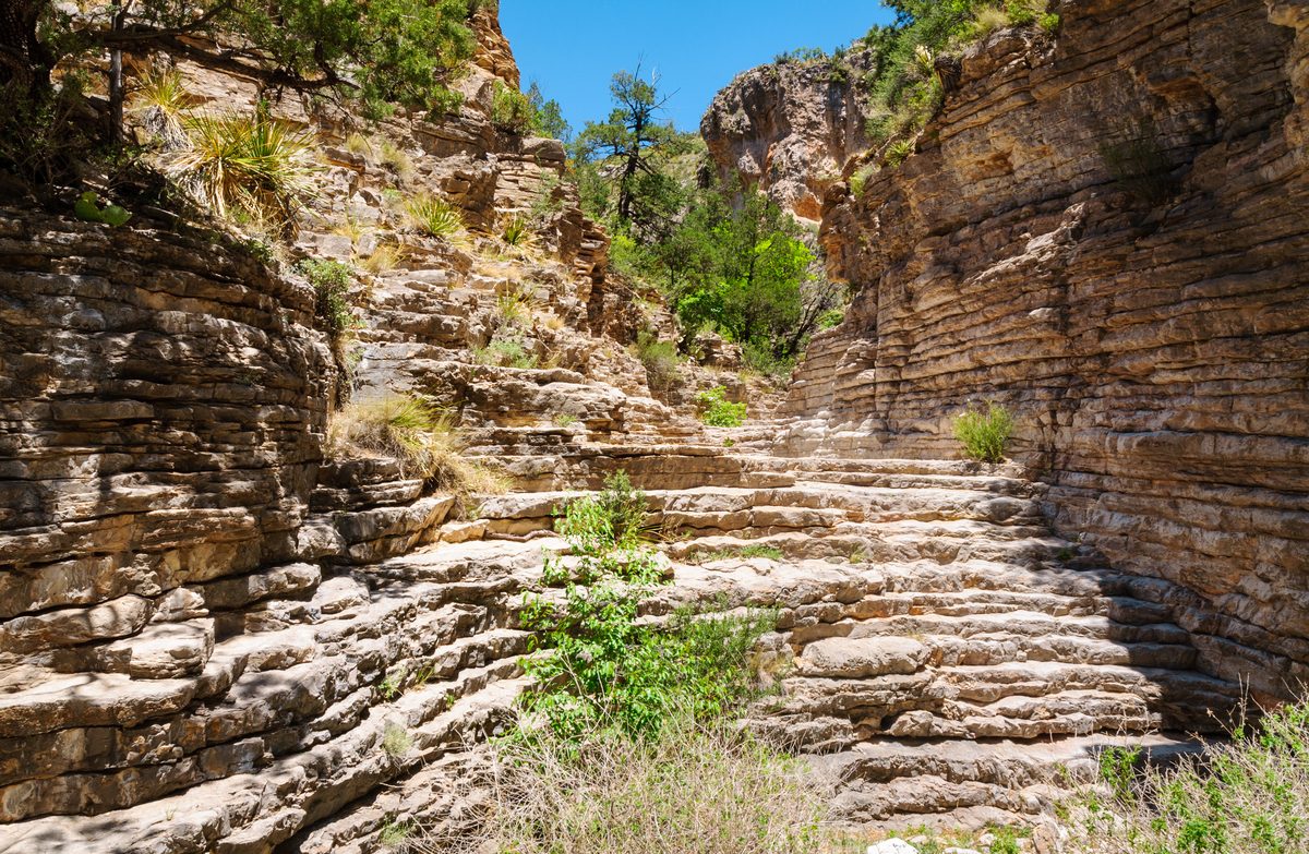 A natural stone staircase of layered limestone formations winds upward through a narrow canyon corridor, flanked by stratified rock walls with desert vegetation. Bright midday sunlight illuminates the warm tan and golden-brown rocks against a vivid…