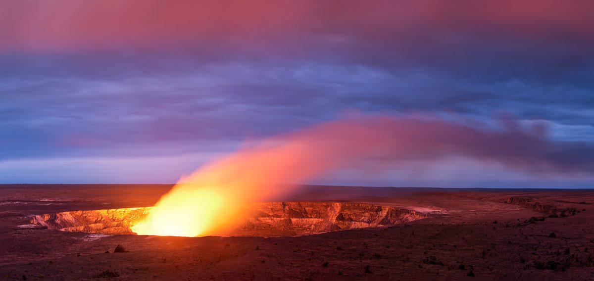 A glowing lava lake inside Halemaʻumaʻu Crater at Kīlauea volcano illuminates the caldera walls with intense orange and yellow light, casting a dramatic plume of volcanic gas across a twilight sky of deep blues and purples.