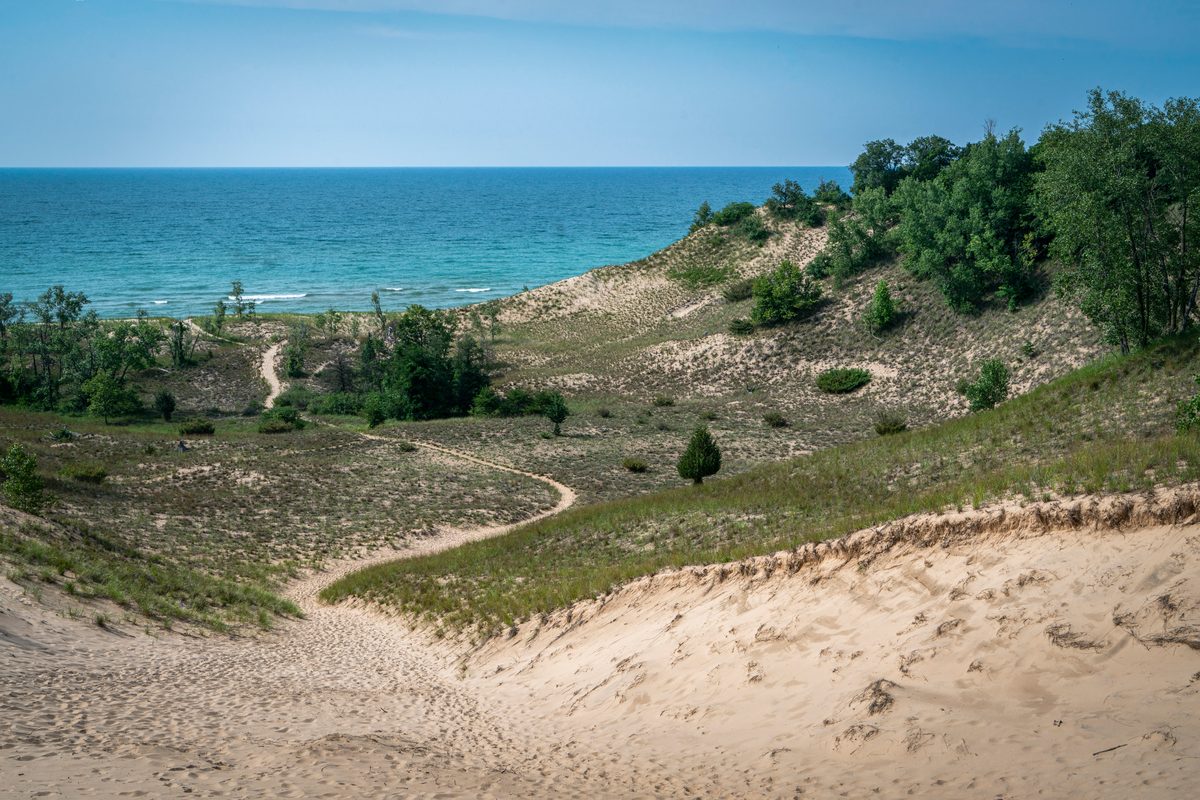 A sweeping view of sandy dunes with sparse vegetation and a winding trail leading down toward the vivid turquoise-blue waters of Lake Michigan, bathed in bright midday summer light with a clear sky.