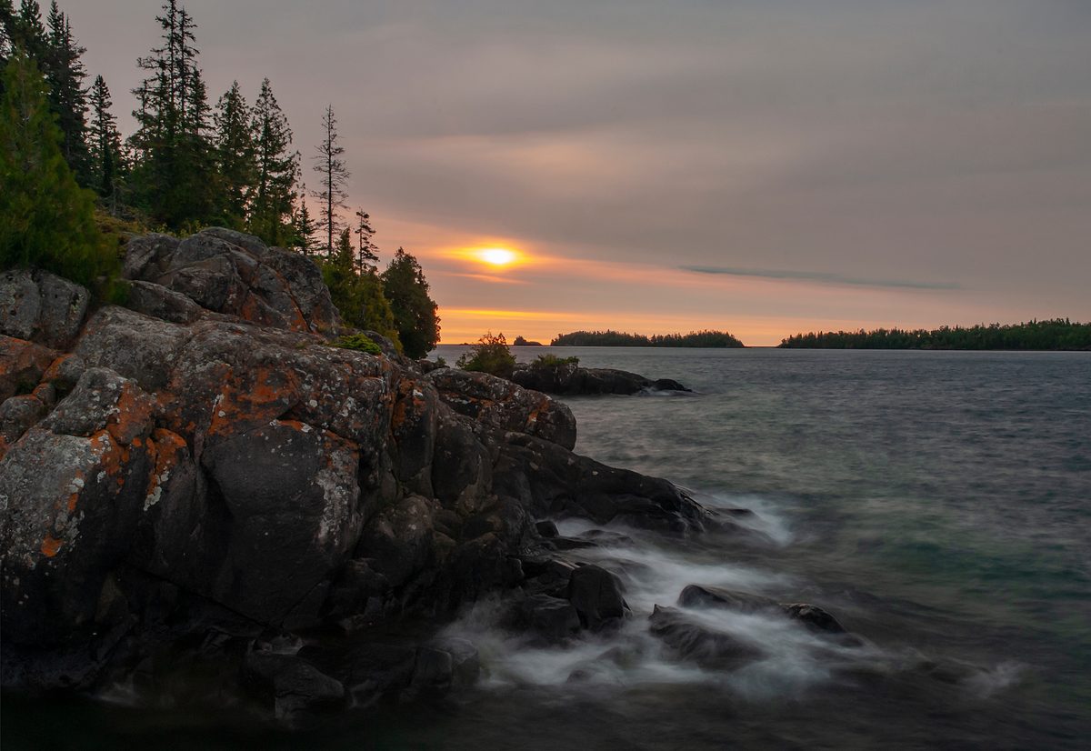A dramatic sunset over a large lake viewed from a rocky, lichen-covered shoreline lined with boreal conifers, with forested islands silhouetted in the distance. The long-exposure capture renders the lapping water silky and ethereal beneath a hazy,…