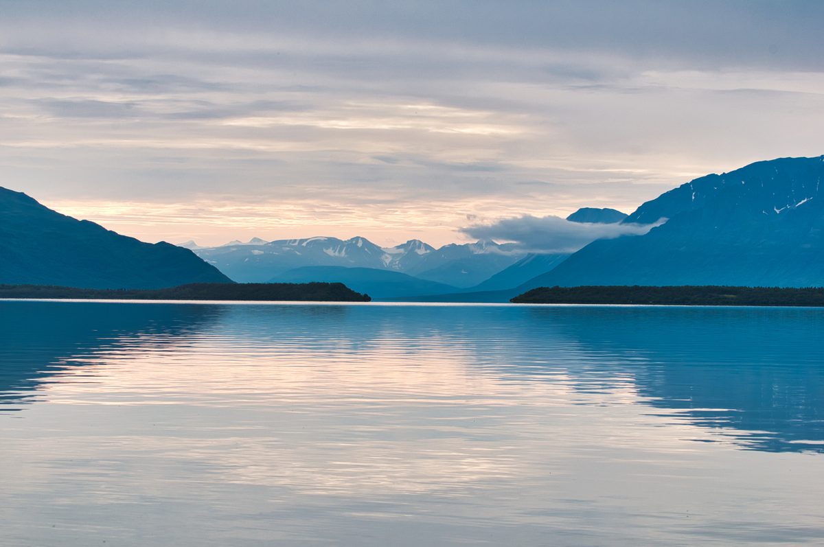 A serene glacial lake reflects soft pastel twilight hues beneath a layered overcast sky, framed by dark silhouetted mountains and snow-capped peaks receding into the misty distance. The calm, mirror-like water and cool blue tones create a tranquil,…