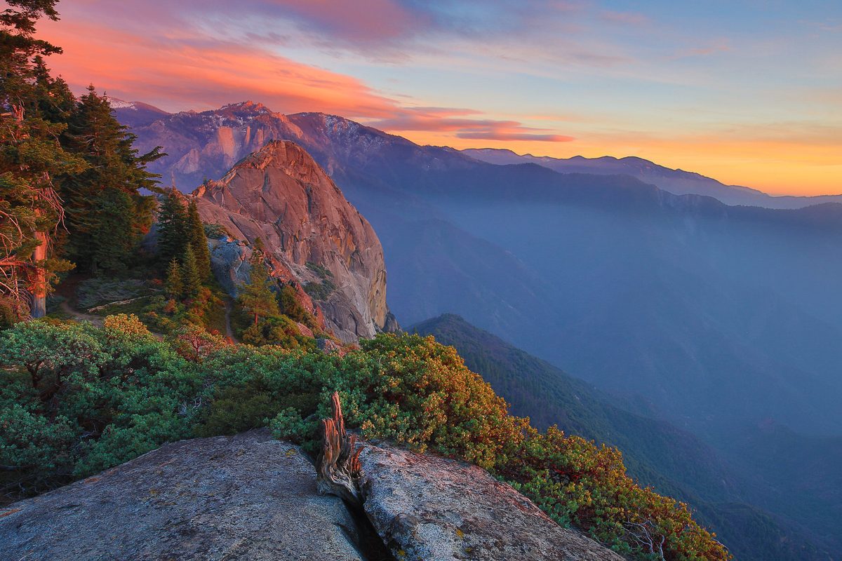 A dramatic sunset view from Moro Rock in Sequoia National Park, showcasing a massive granite dome flanked by conifers and manzanita shrubs, with layered misty mountain ridges receding into the distance under a vivid orange and pink sky.