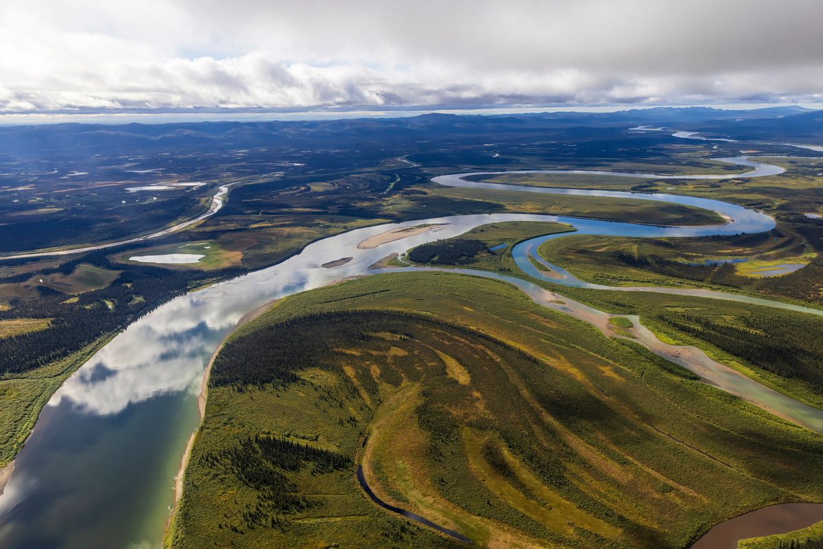 An aerial view of a vast braided and meandering river system winding through boreal forest and tundra flats, likely the Yukon or Kobuk River in Alaska, under dramatic partly cloudy skies with soft diffused light reflecting off the calm water.
