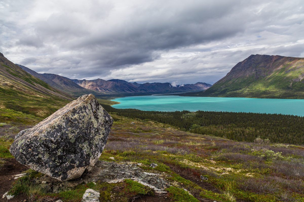 A striking glacial lake with vivid turquoise water nestled between rugged mountain ranges, framed by a large lichen-covered boulder in the foreground and a dense boreal forest below under dramatic overcast skies.