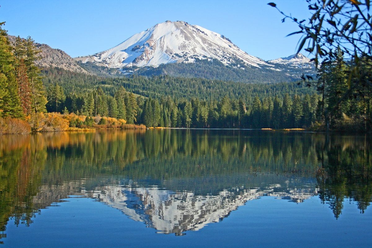 A snow-capped volcanic peak — likely Lassen Peak in Lassen Volcanic National Park, California — rises above a dense evergreen forest, with its reflection mirrored perfectly in the calm, glassy surface of Manzanita Lake. The autumn foliage adds warm…