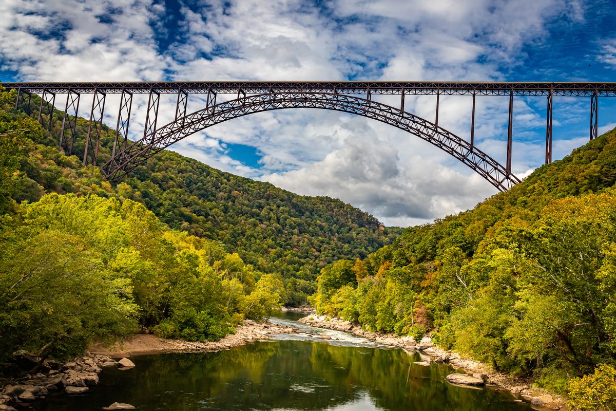 The iconic New River Gorge Bridge, a massive steel arch bridge spanning the densely forested gorge above the New River in West Virginia, photographed from below with lush green canopy and partly cloudy blue skies creating a dramatic, majestic mood.