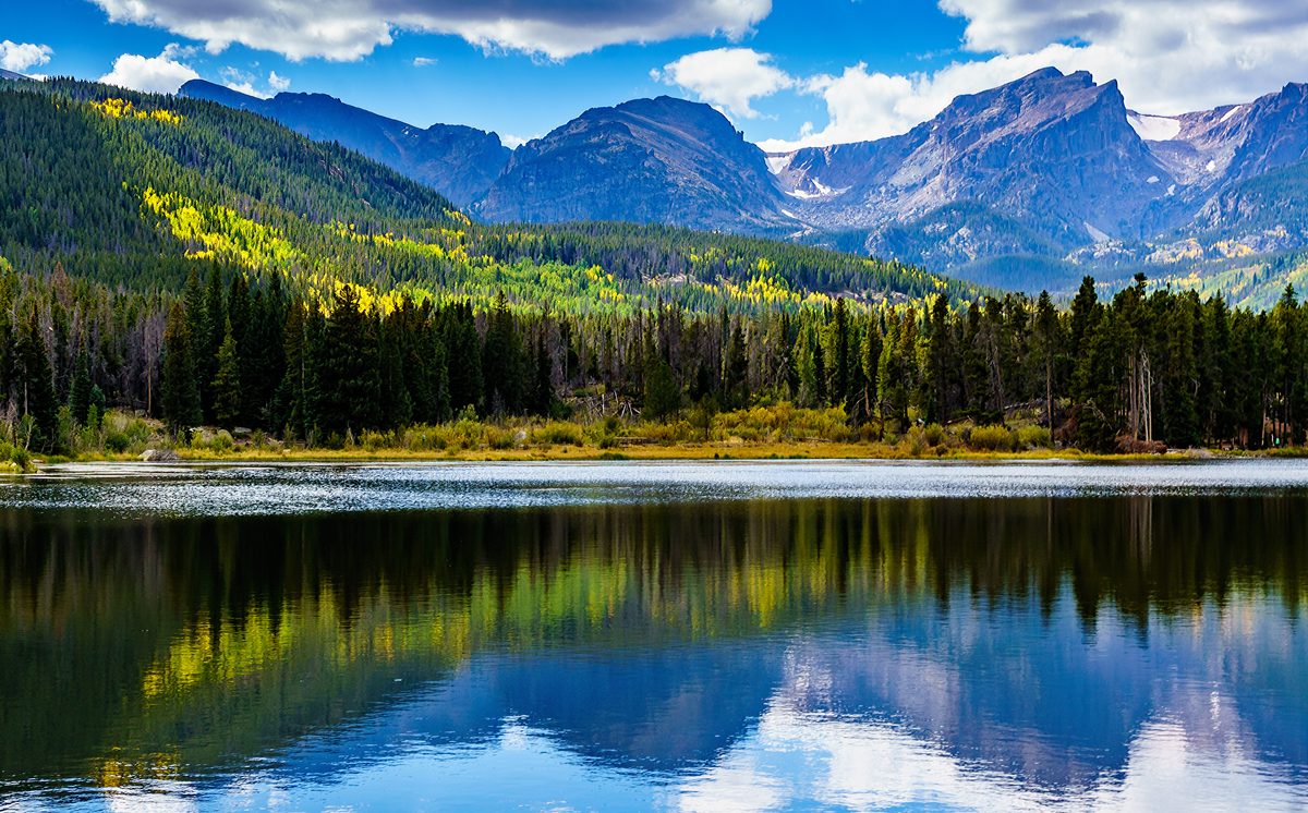 A serene alpine lake in Rocky Mountain National Park reflects the surrounding evergreen forest and rugged mountain peaks under a partly cloudy blue sky. The scene captures early autumn foliage with golden aspen trees dotting the hillsides, creating…