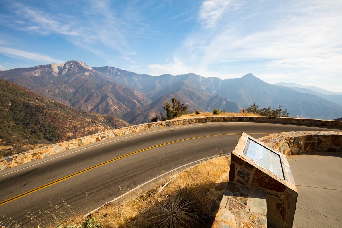 A winding mountain road curves along a stone-walled scenic overlook with a wayside exhibit panel, offering a sweeping view of rugged Sierra Nevada peaks and canyon ridgelines under a bright blue sky with wispy clouds.