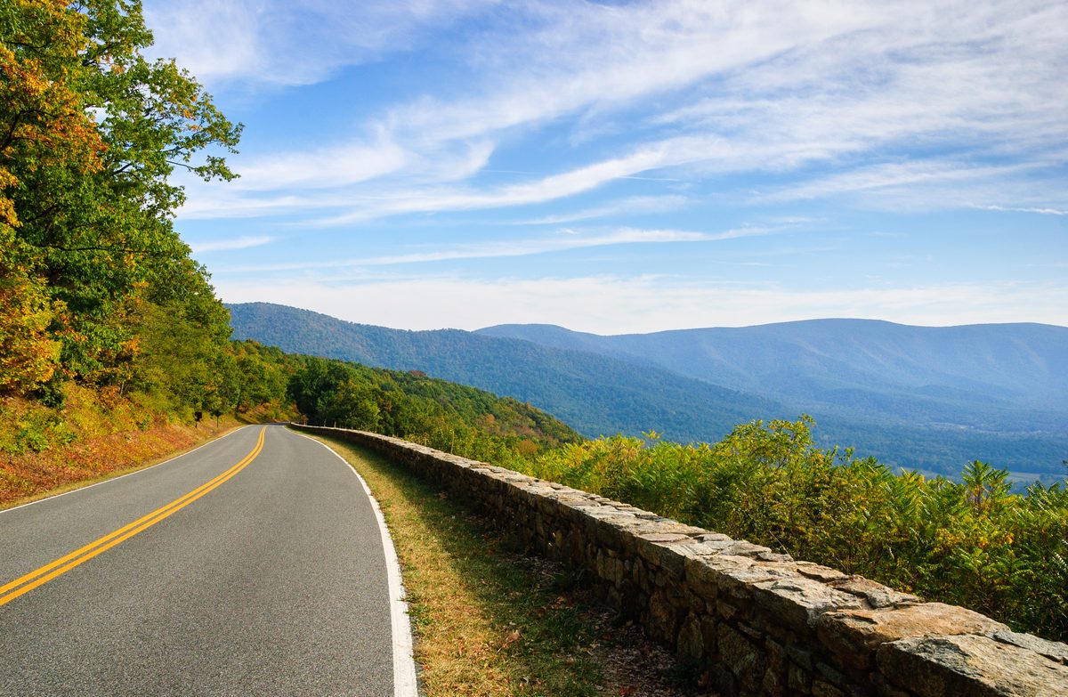 A scenic two-lane mountain road with a rustic stone guardrail curves along a ridgeline in Shenandoah National Park, with sweeping views of the Blue Ridge Mountains layered in soft haze under a bright autumn sky.
