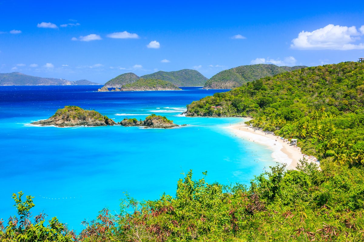 A stunning aerial view of Trunk Bay in St. John, US Virgin Islands National Park, featuring vivid turquoise and deep blue waters curving around a white sandy beach flanked by lush tropical hillsides. The scene is bathed in brilliant midday sunlight,…