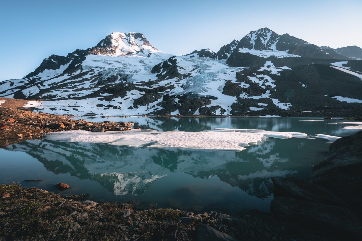 A glacially-fed alpine lake reflects snow-capped mountain peaks and a glacier in still, turquoise waters, with floating ice floes in the foreground and warm early-morning or late-evening light kissing the rocky shoreline.
