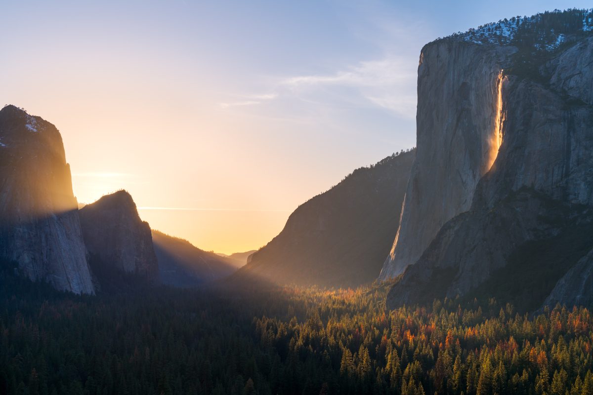 Yosemite Valley at golden hour featuring El Capitan's sheer granite face with Horsetail Fall glowing orange in the famous 'Firefall' effect, framed by forested valley floor with autumn foliage and a warm sunset illuminating misty air between canyon…