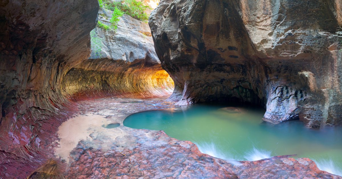 A stunning slot canyon interior known as 'The Subway' in Zion National Park, featuring smooth water-carved sandstone walls glowing with warm amber and rust tones, framing a serene turquoise pool below. The ethereal lighting filters through the…